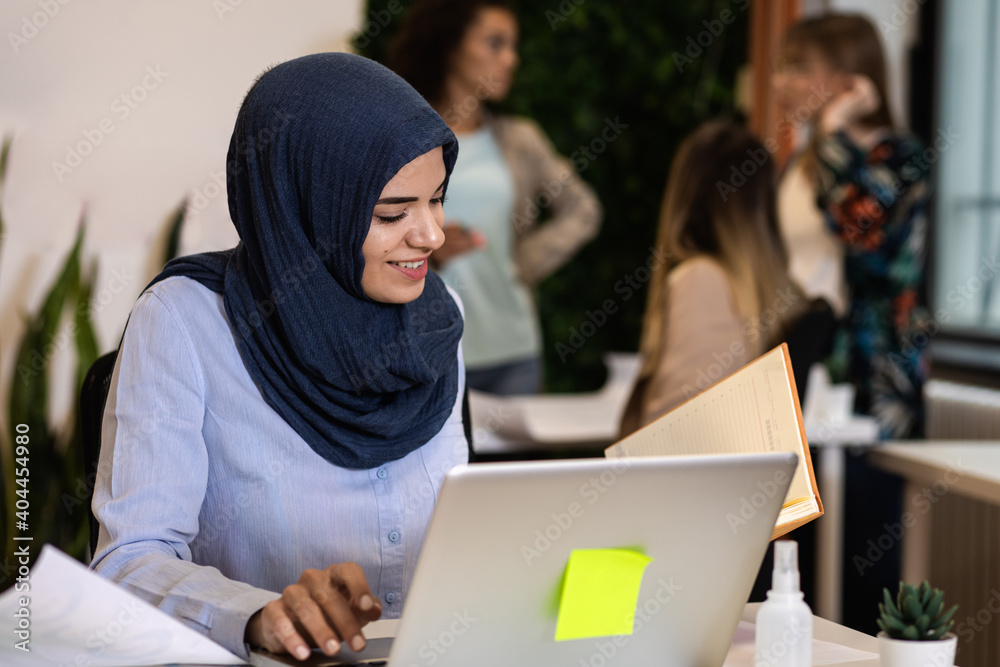Portrait of a young Muslim businesswoman working in an office while sitting at a table with colleagues in the background. Portrait of a young Muslim businesswoman working in an office while sitting at a table with colleagues in the background.