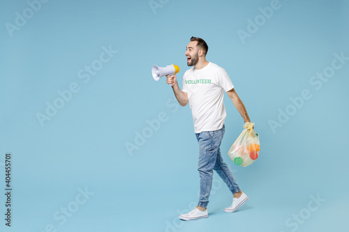 Full length of funny young man in white volunteer t-shirt jumping hold trash bag scream in megaphone isolated on blue background studio. Voluntary free assistance help trash sorting recycling concept.