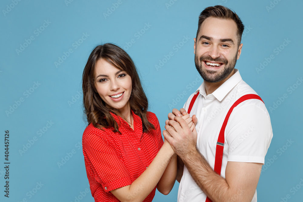 Side view of smiling young couple two friends man woman wearing white red clothes holding hands looking camera isolated on blue color background studio portrait. St. Valentine's Day holiday concept. Side view of smiling young couple two friends man woman wearing white red clothes holding hands looking camera isolated on blue color background studio portrait. St. Valentine's Day holiday concept.