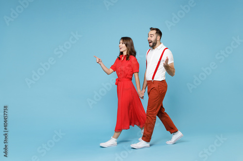 Full length of shocked young couple two friends man woman in white red clothes pointing index finger aside spreading hands isolated on blue background studio. St. Valentine's Day holiday concept.