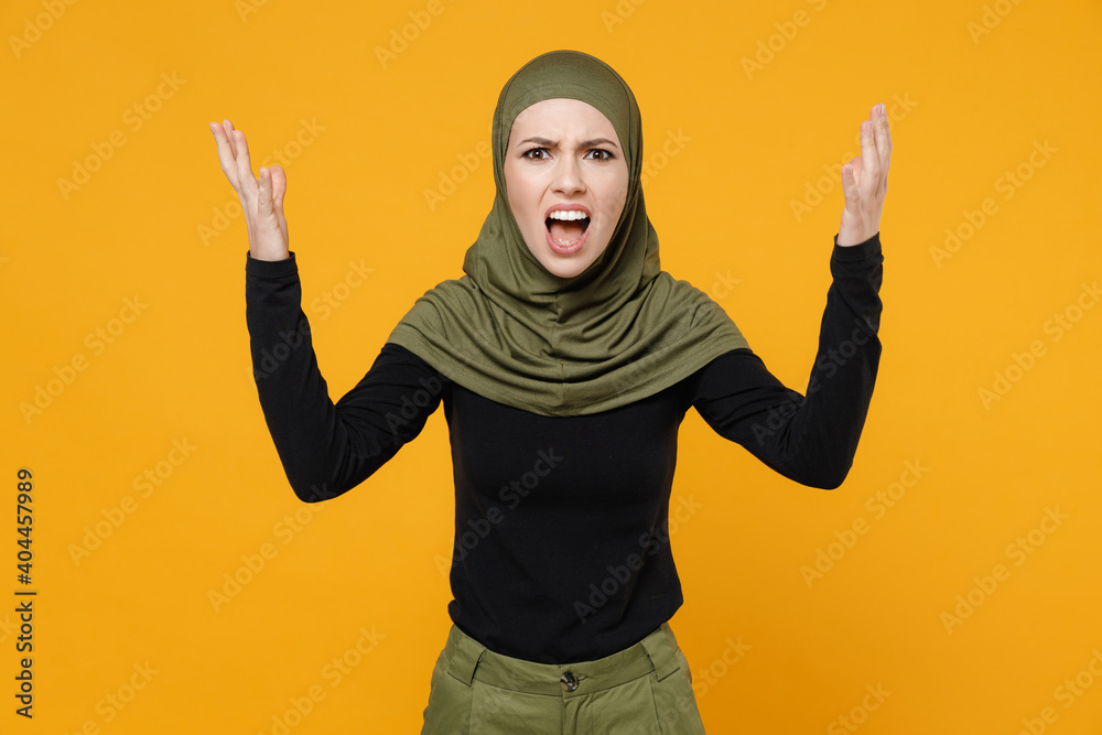Angry irritated young arabian muslim woman in hijab black green clothes spreading hands screaming swearing isolated on yellow color background, studio portrait. People religious lifestyle concept.