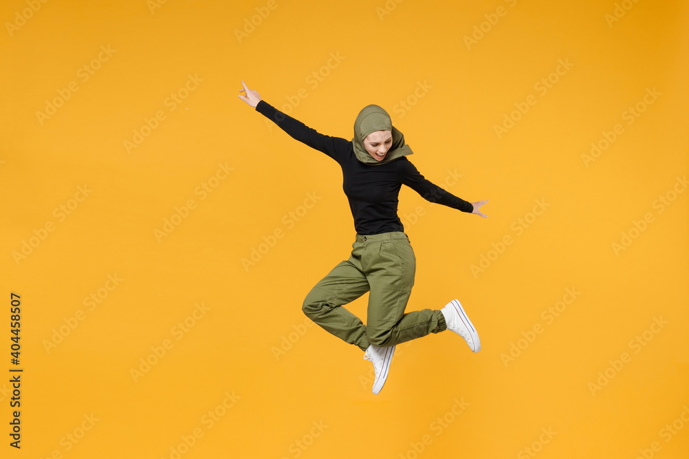 Fototapeta premium Full length of cheerful young arabian muslim woman wearing hijab black green clothes jumping spreading hands isolated on yellow color background studio portrait. People religious lifestyle concept.