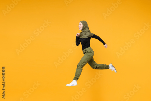 Full length side view of cheerful young arabian muslim woman in hijab black green clothes jumping like running isolated on yellow color background studio portrait. People religious lifestyle concept.