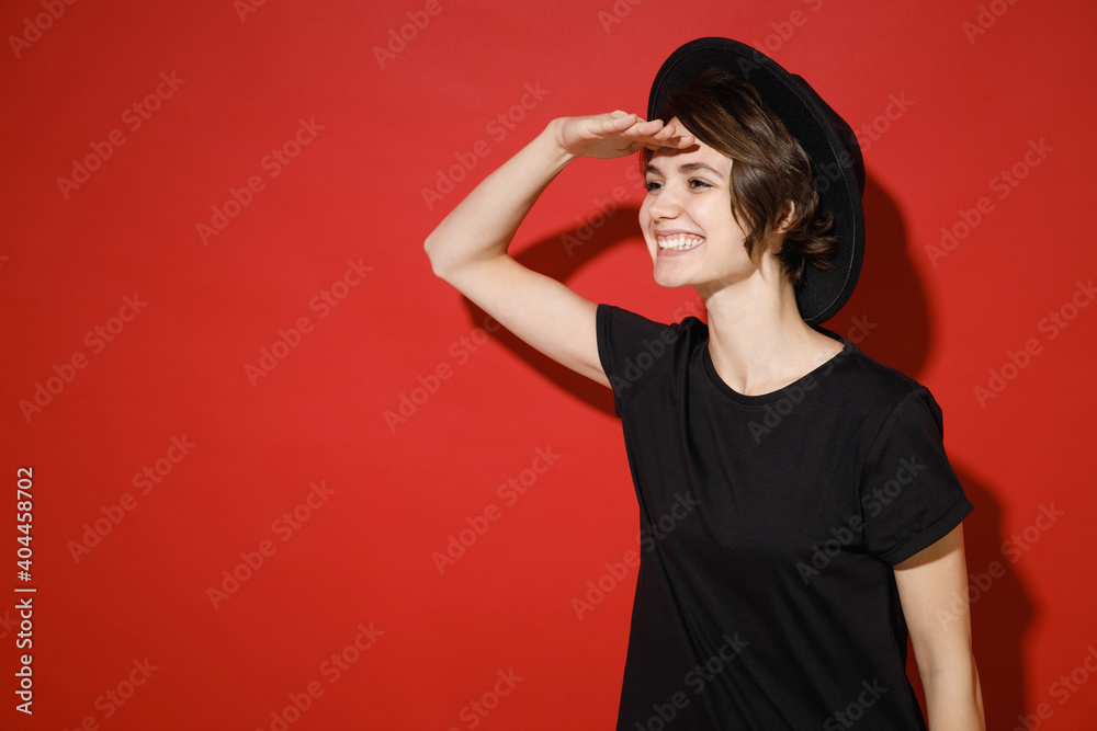 Smiling young brunette woman 20s years old wearing casual basic black t-shirt hat standing holding hand at forehead looking far away distance isolated on bright red color background studio portrait. Smiling young brunette woman 20s years old wearing casual basic black t-shirt hat standing holding hand at forehead looking far away distance isolated on bright red color background studio portrait.