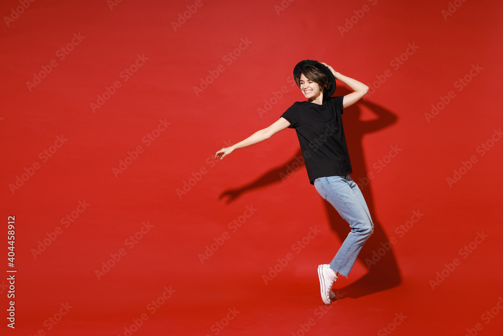 Full length side view of cheerful smiling young brunette woman 20s in casual basic black t-shirt hat standing on toes dancing put hand on head isolated on bright red color background studio portrait.