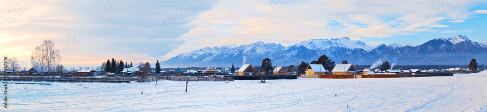 Panoramic view of the Tunka foothill valley with the village against the background of snow-capped mountains at sunset on a cold winter day. Siberia, Buryatia, Tunka, Arshan, Eastern Sayan Mountains