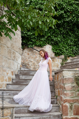 Beautiful woman with purple hair in a white wedding dress walks in the garden. Natural makeup