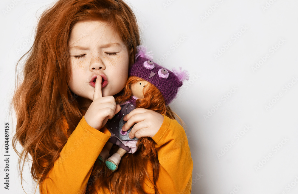 Cheerful small red haired girl in yellow comfortable longsleeve standing holding toy doll in hands and showing silence sign with finger over white background, copy space. Happy childhood, stylish look