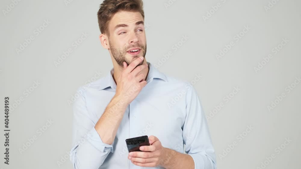 A thinking young businessman is chatting using his smartphone standing isolated over a grey background in the studio A thinking young businessman is chatting using his smartphone standing isolated over a grey background in the studio