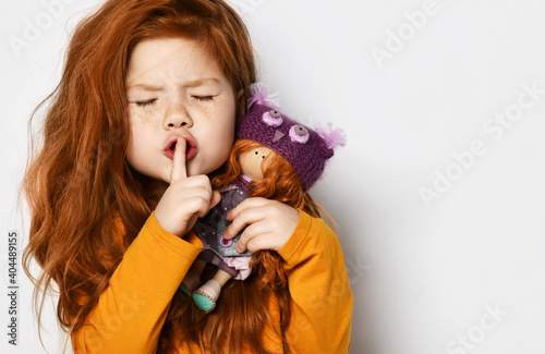 Obraz na płótnie Cheerful small red haired girl in yellow comfortable longsleeve standing holding toy doll in hands and showing silence sign with finger over white background, copy space. Happy childhood, stylish look w salonie