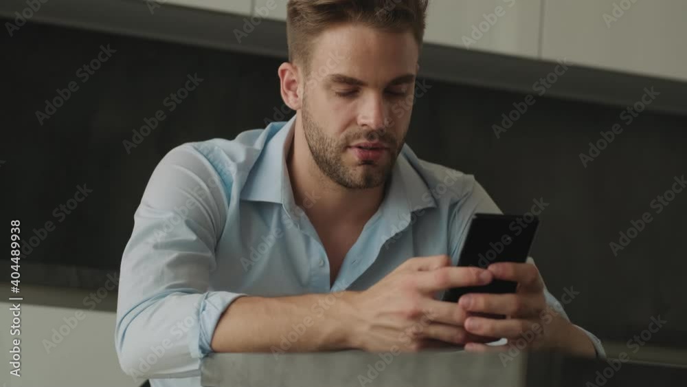 A calm young businessman is typing on his smartphone sitting in the kitchen at home A calm young businessman is typing on his smartphone sitting in the kitchen at home