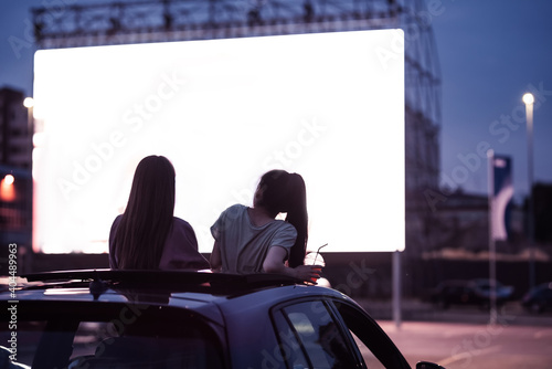 Rear view of two female friends sitting in the car while watching a movie in an open air cinema with a big white screen photo