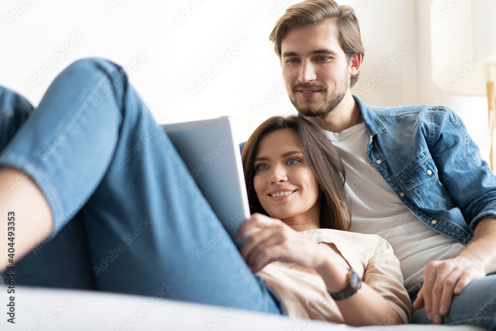 © opolja - Young couple sitting on couch at home, using a tablet PC for Internet and social media.