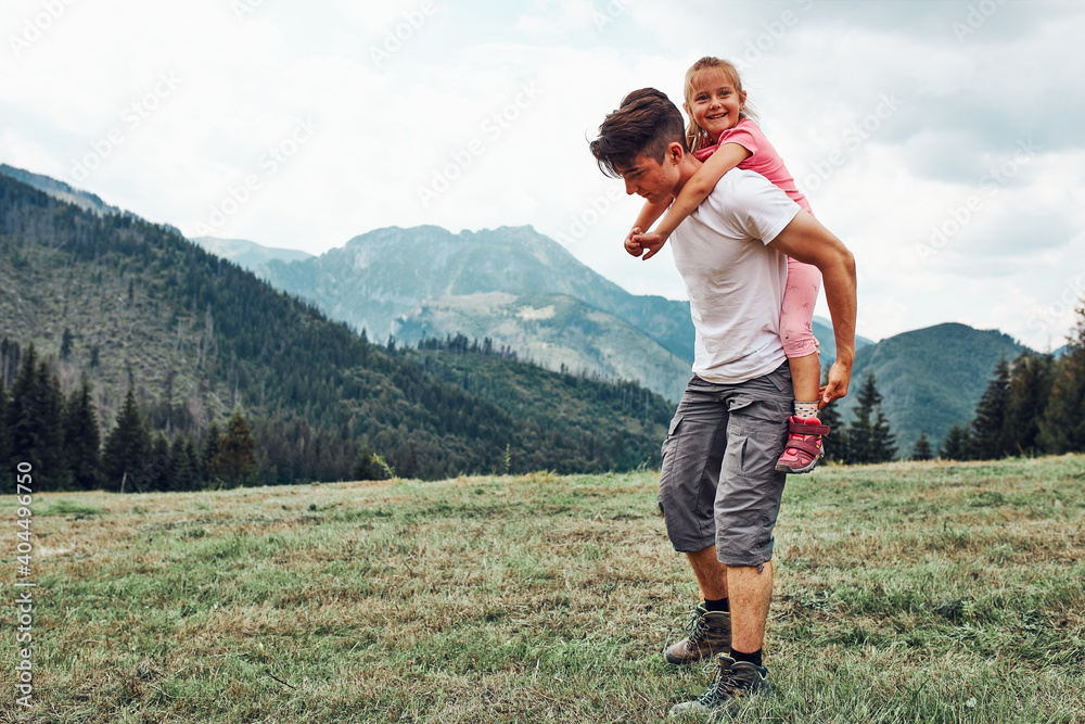 Young man holding little girl on his back. Child playing with her elder brother riding on his back enjoying summer day together. Happy siblings playing in the field during vacation trip in mountains
