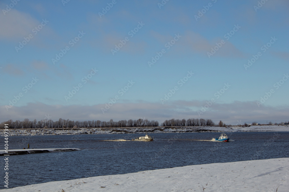 Two ships sail along the channel of the northern river. At the beginning of winter, two ships sail along a narrow channel among the banks covered with snow.