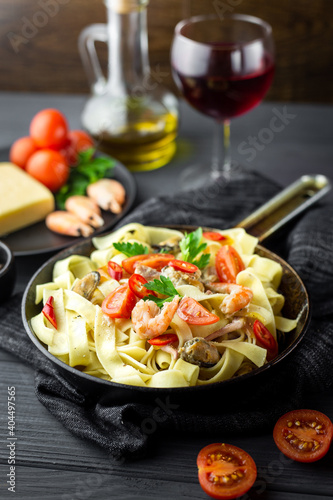 Small pan with fettuccine and sea food decorated with greens and cherry tomatoes, black napkin, a glass of wine, bottle with olive oil around on the black wooden table. Close up