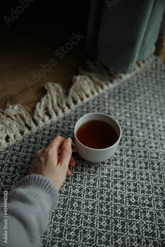 female hand in a gray sweater holds a white mug of tea on the background of a vintage carpet