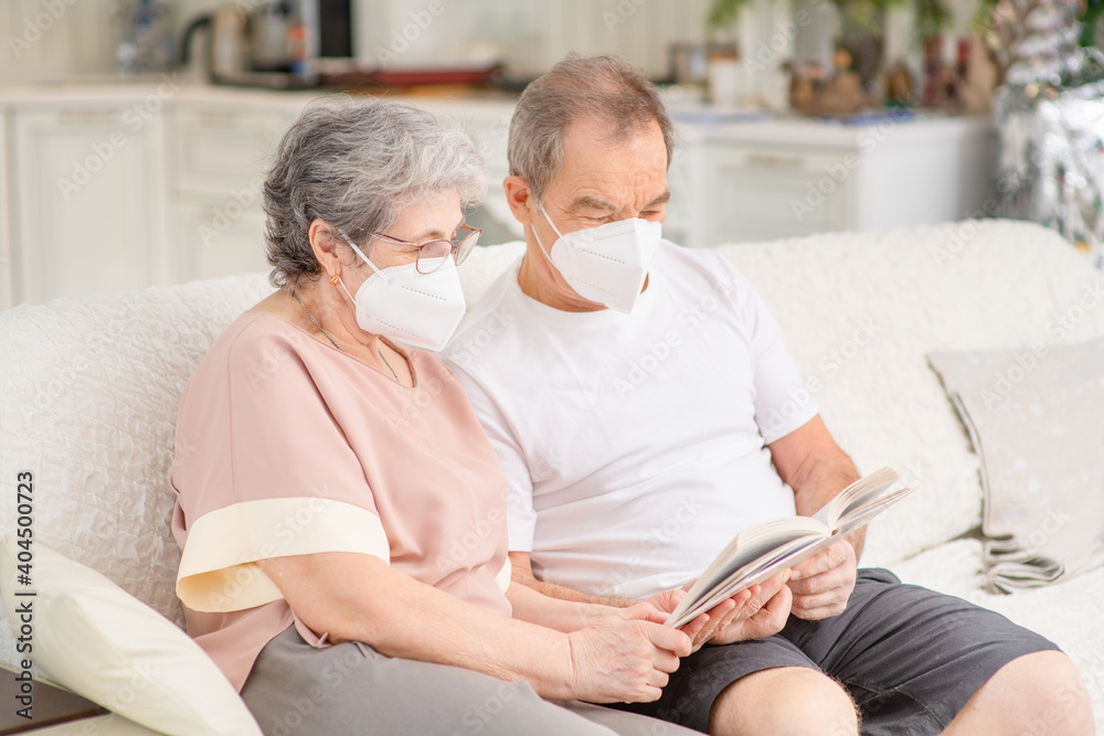 An elderly couple with medical masks are sitting on the couch at home and reading a book. Ways not to get bored during lockdown and develop while sitting at home. Quarantine during coronavirus pandemi An elderly couple with medical masks are sitting on the couch at home and reading a book. Ways not to get bored during lockdown and develop while sitting at home. Quarantine during coronavirus pandemi