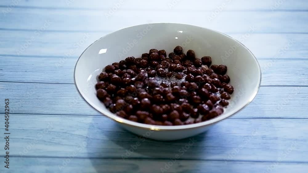 White plate of ready made breakfast cereal on blue wooden table. Pour chocolate crunchy balls with milk. Slow motion video of cooking fast food. White plate of ready made breakfast cereal on blue wooden table. Pour chocolate crunchy balls with milk. Slow motion video of cooking fast food.
