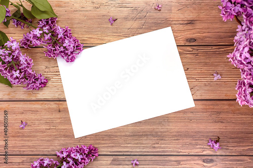 White blank sheet of paper with blooming lilac flowers on wooden background. Invitation or greeting card for Valentine's Day and Mother's Day. Top view, flat lay, mock up, copy space. Spring concept.