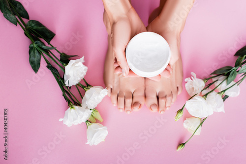top view of female hands with cosmetic cream near feet and white eustoma flowers on pink background