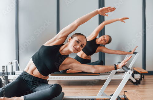 With help of gym equipment. Two women in sportive wear and with slim bodies have fitness yoga day indoors together