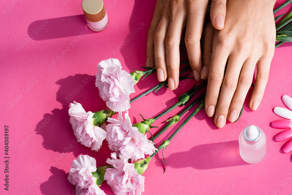 top view of female hands near carnation flowers, palette of fake nails, vials with cuticle remover and nail varnish on pink background