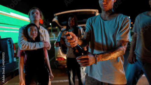 A group of well-dressed friends waiting for a young tattooed guy to open a bottle of champagne during a party with a car and a big led screen on a background