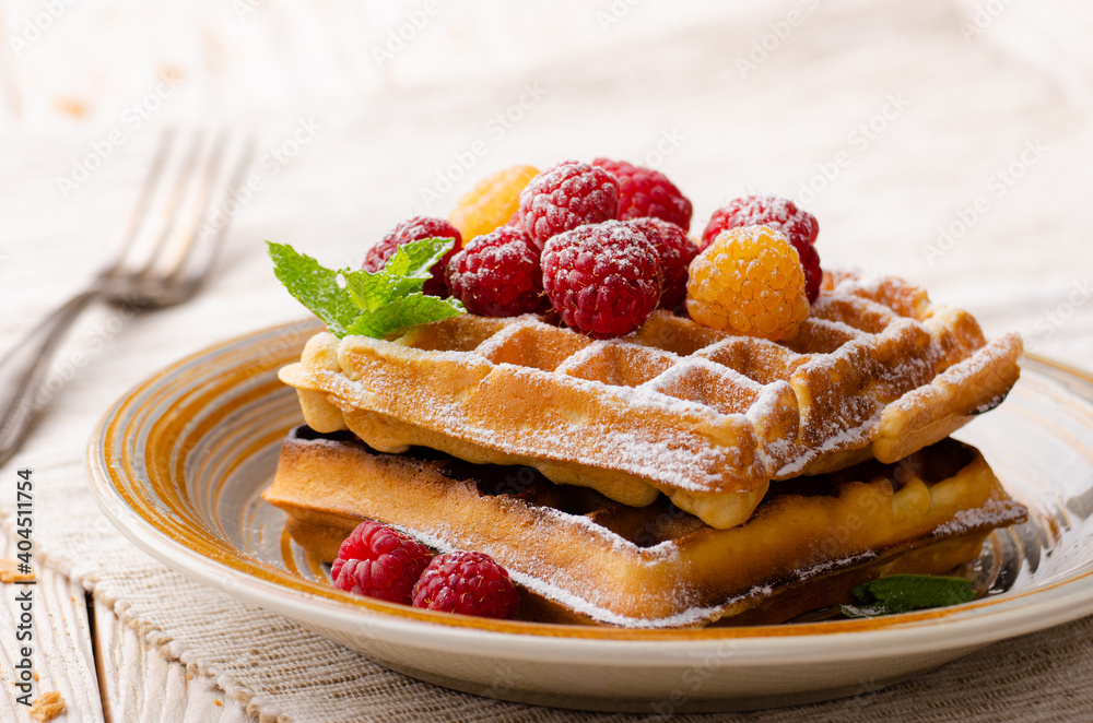 Belgian waffles served with raspberries and mint leaf dusted with powdered sugar on white wooden kitchen table Belgian waffles served with raspberries and mint leaf dusted with powdered sugar on white wooden kitchen table