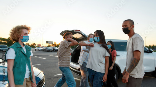 Young casually dressed friends of different nationalities greeting each other with elbows wearing masks standing outside on a parking site with their cars on a background