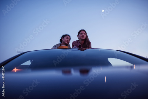 Two girls lying on the roof of a car with popcorn bags and drinks while watching a movie in an open air cinema in the evening © Svitlana
