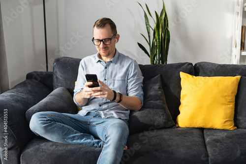 A handsome guy wearing stylish glasses using smartphone for remote work, answering email, texting messages sitting at contemporary designed home on the comfortable sofa