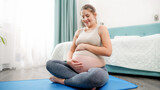 Beautiful smiling pregnatn woman in sportrs outfit sitting on fitness mat and stroking her big belly. Concept of healthy lifestyle, healthcare and sports during pregnancy