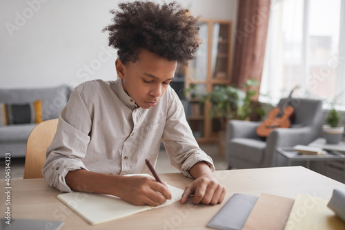 Portrait of teenage African-American boy doing homework and writing in notebook while sitting at desk in home interior, copy space