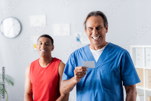 happy doctor holding blank card near african american patient in sportswear on blurred background