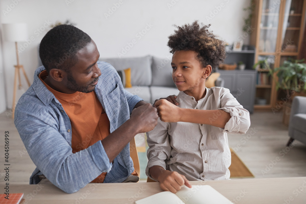 Portrait of African-American father fist bumping smiling son while doing homework together at home - obrazy, fototapety, plakaty 