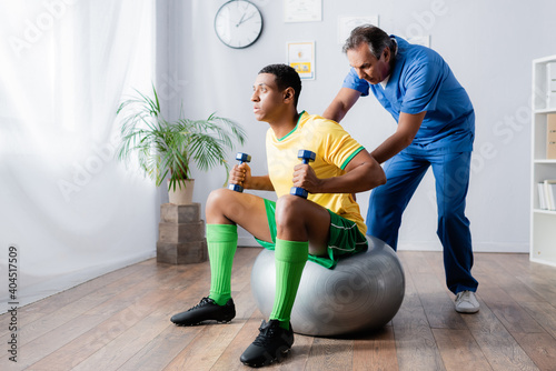 african american sportsman working out with dumbbells on fitness ball near physiotherapist photo