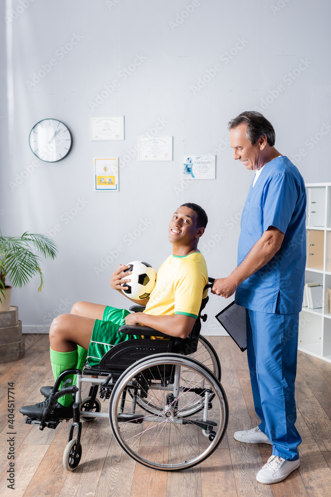 cheerful african american sportsman in wheelchair holding football and smiling near doctor cheerful african american sportsman in wheelchair holding football and smiling near doctor