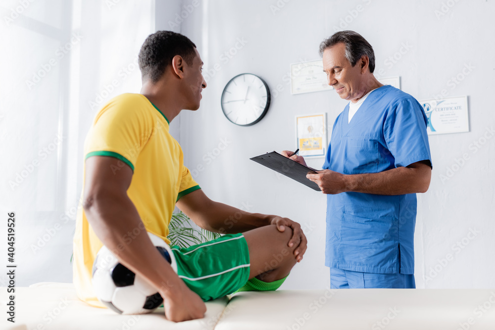 Obraz premium doctor writing prescription near smiling african american football player sitting with ball on massage table