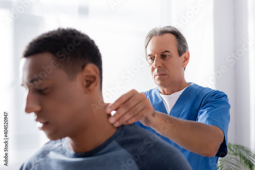 masseur massaging injured neck of african american patient on blurred foreground