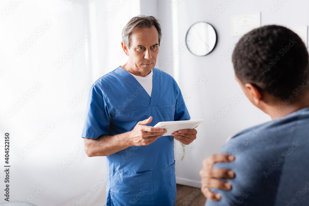 mature doctor holding digital tablet near african american patient on blurred foreground mature doctor holding digital tablet near african american patient on blurred foreground