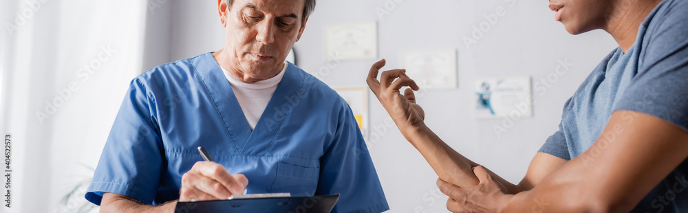 mature doctor writing prescription on clipboard near injured african american man, banner