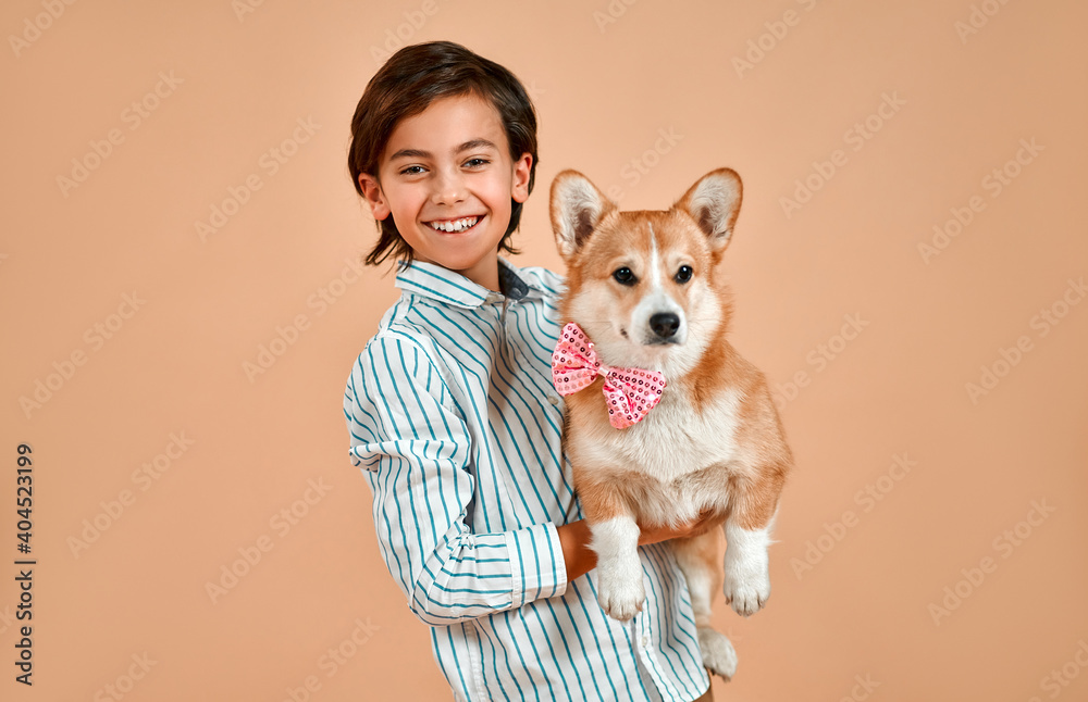 Cute smiling boy in a striped shirt holds a dog with a bow on his neck isolated on a powdery peach background.Valentine's Day.