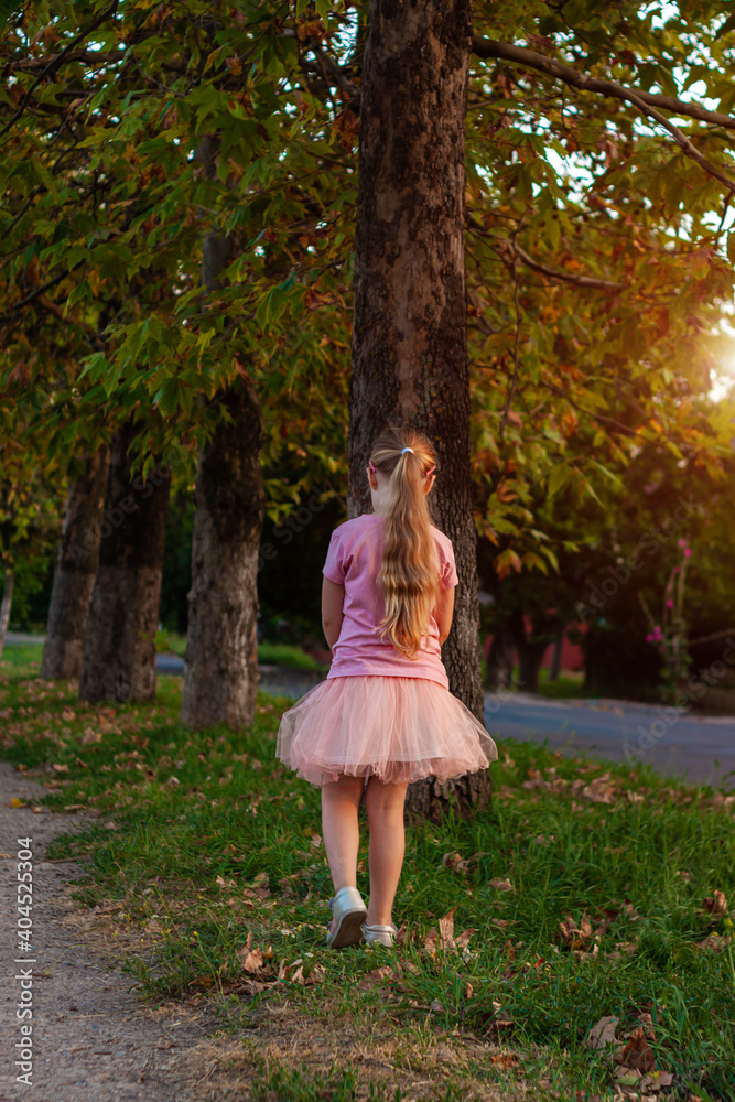 Cheerful little girl with long blonde hair in pink tulle skirt walking alone on empty street on sunset. Greeting card. Cheerful little girl with long blonde hair in pink tulle skirt walking alone on empty street on sunset. Greeting card.