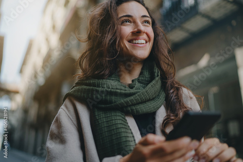 Young attractive smiling broadly woman wearing light-colored coat and green scarf, walking city streets on a sunny day and using smartphone for communication. Good vibes. Modern technology advantages photo