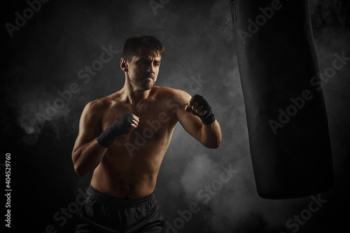 sporty shirtless boxer in black boxing wraps punching in boxing bag on dark background with smoke © producer