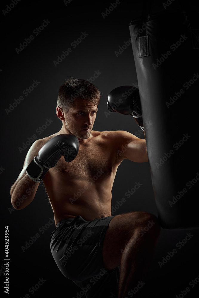 muscular boxer in black boxing gloves punching in boxing bag on dark background