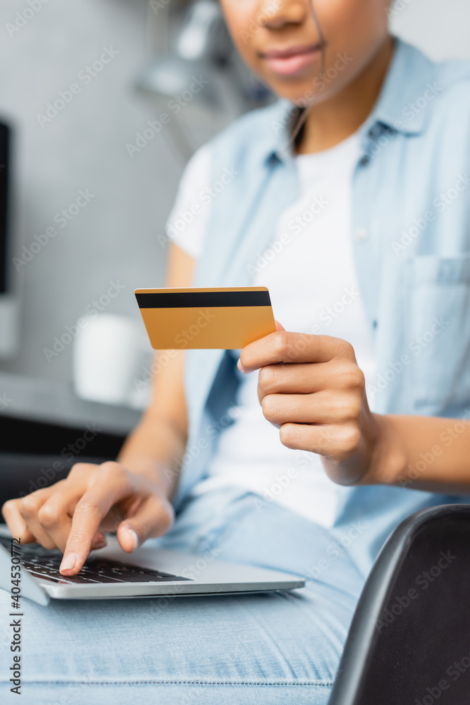 partial view of african american woman holding credit card while using laptop, blurred foreground partial view of african american woman holding credit card while using laptop, blurred foreground
