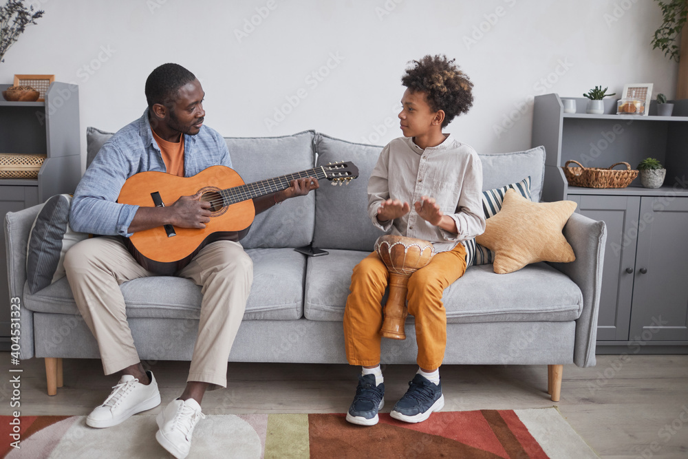Horizontal full shot of African American man and his son spending time together at home playing guitar and djembe