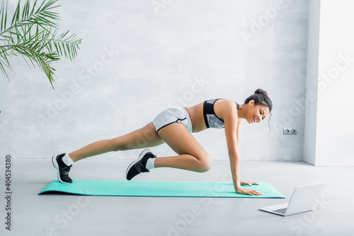 side view of happy african american sportswoman practicing one legged plank pose near laptop